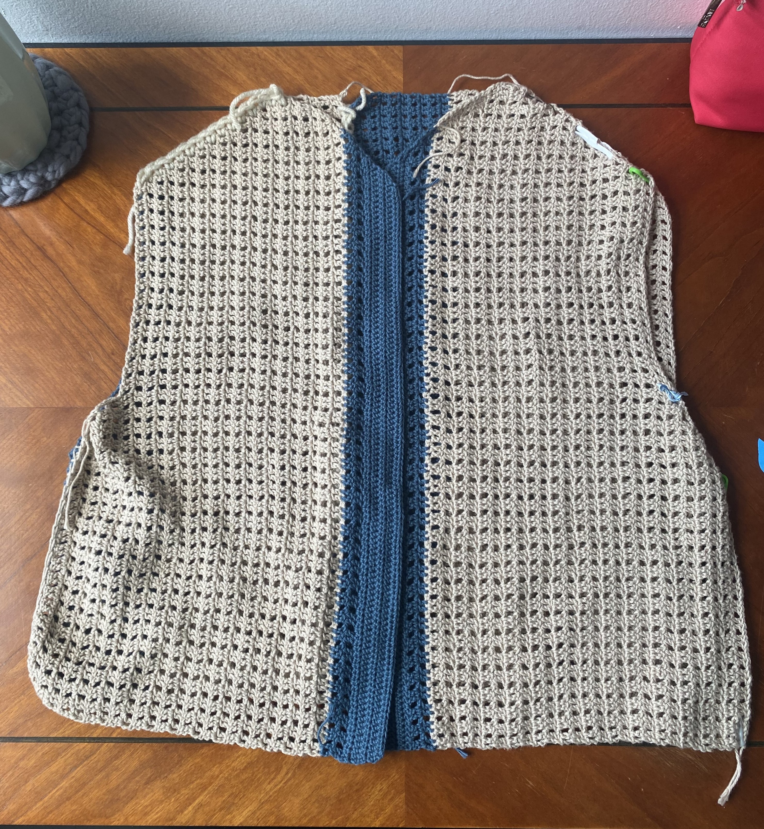 a unfinished crochet mesh vest in beige with a blue lining with some crochet markers holding it together